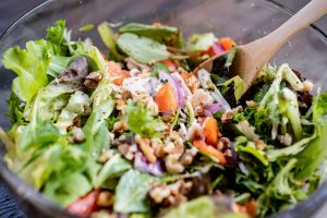 Close-up of a fresh mixed green salad with walnuts, tomatoes, and a wooden spatula in a glass bowl.